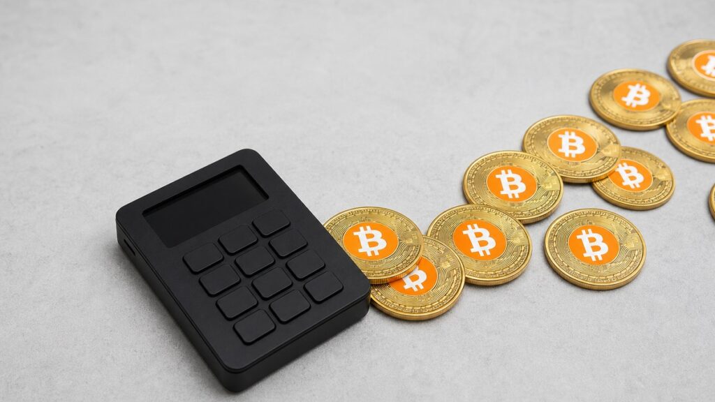 Eine schwarze Hardware-Wallet auf einer hellen Steinfläche, von der eine Spur goldener Bitcoin-Münzen nach rechts aus dem Bild gleitet.