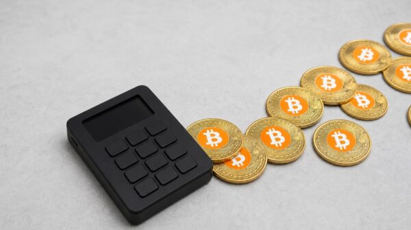 Eine schwarze Hardware-Wallet auf einer hellen Steinfläche, von der eine Spur goldener Bitcoin-Münzen nach rechts aus dem Bild gleitet.