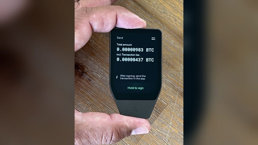 Zwei Hände halten eine schwarze Hardware-Wallet, auf deren Bildschirm eine Bitcoin-Überweisung zur Bestätigung angezeigt wird