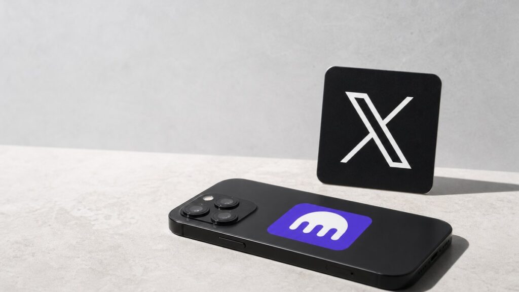 Ein Smartphone liegt mit der Rückseite nach oben auf hellem Stein, darauf das violette Kraken-Zeichen mit dem weißen Tintenfisch; dahinter steht ein schwarzer Aufsteller mit dem weißen X-Logo.