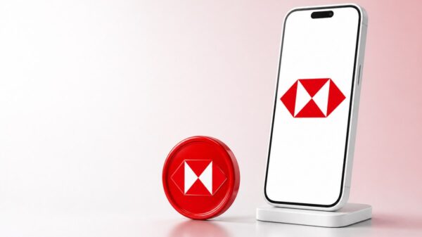 Ein weißes Smartphone auf einem Ständer zeigt das rot-weiße HSBC-Sechseck, daneben steht eine glänzende rote Münze mit demselben Zeichen.