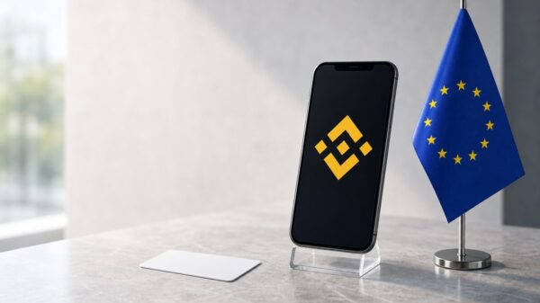 Ein Smartphone auf einem durchsichtigen Ständer zeigt das gelbe Binance-Zeichen, daneben steht eine kleine EU-Tischflagge, davor liegt eine weiße Karte.